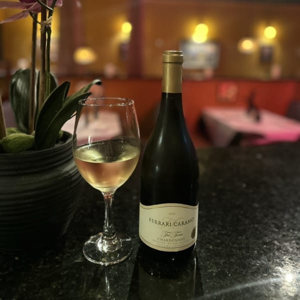A glass of white wine and a bottle of Ferrari Carano Chardonnay on a dark bar top.