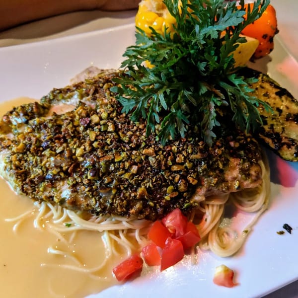 Pistachio-crusted fish over pasta with a creamy sauce, diced tomatoes, and grilled vegetables.