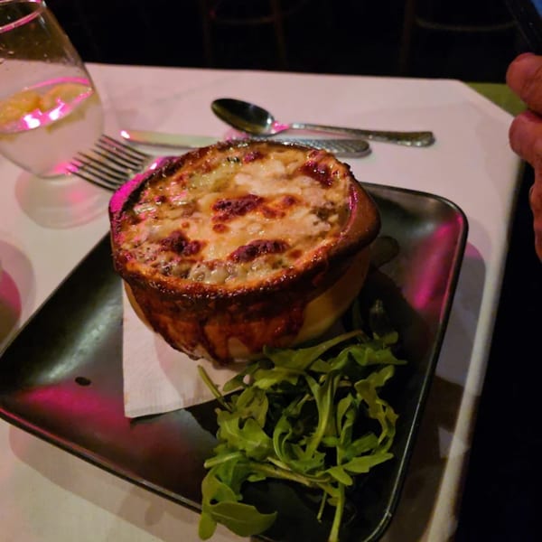 A bowl of French onion soup with melted cheese and a side of greens on a black tray.