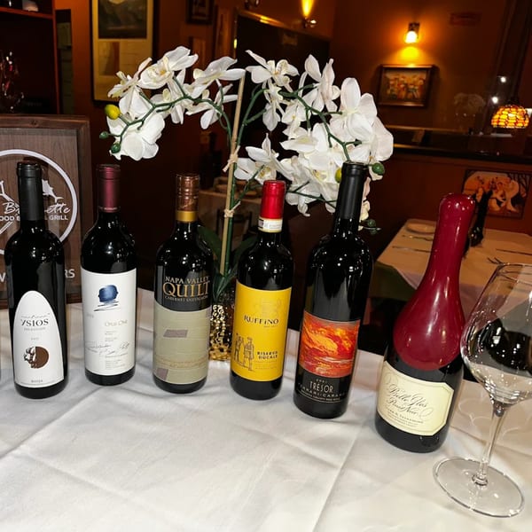 Six bottles of red wine and a wine glass on a white tablecloth with a floral centerpiece.