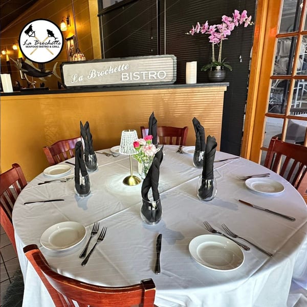 A round dining table set for six with white tablecloths, black napkins, and a floral centerpiece at La Brochette Bistro.