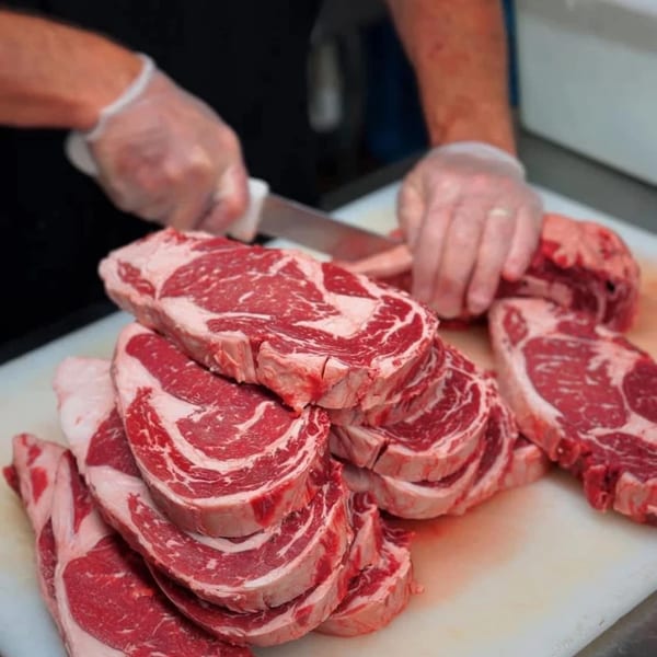 A chef in gloves slices raw ribeye steaks on a white cutting board at La Brochette Bistro.