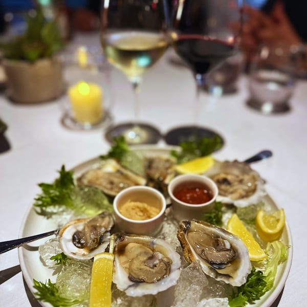 A plate of fresh oysters on ice with lemon wedges and two sauces, accompanied by wine glasses and a candle.