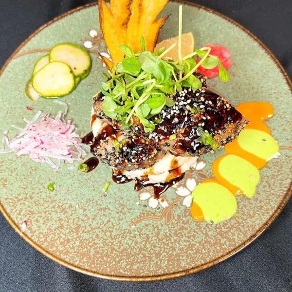 Seared tuna with sesame seeds, greens, sauces, and pickled vegetables on a decorative plate.