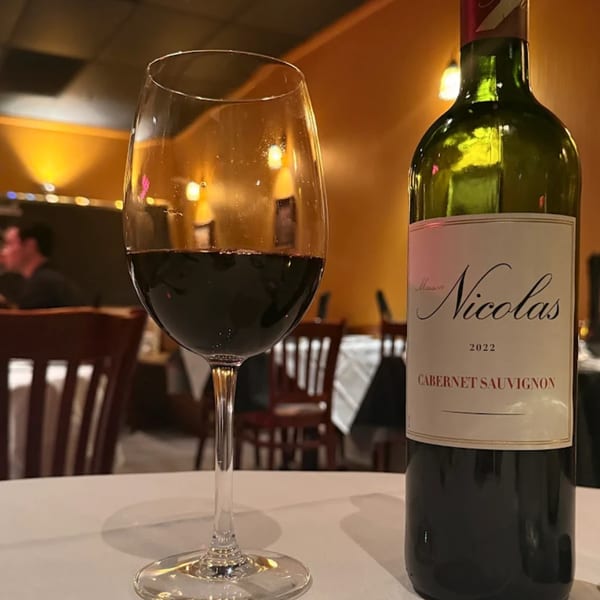 A glass of red wine and a bottle of Maison Nicolas Cabernet Sauvignon on a white tablecloth.