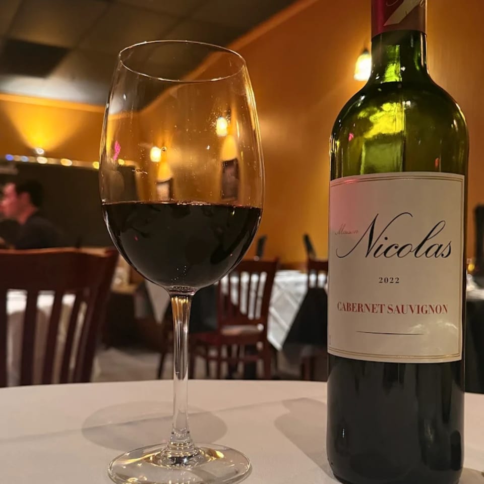 A glass of red wine and a bottle of Maison Nicolas Cabernet Sauvignon on a white tablecloth.