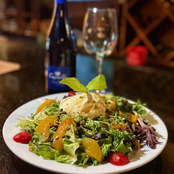 A fresh La Brochette Bistro salad with burrata, peaches, pistachios, and cherry tomatoes, with wine in background.