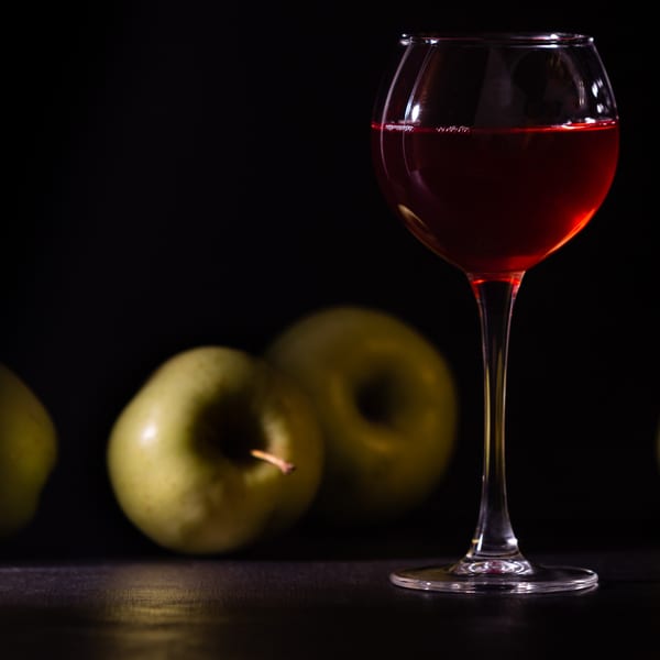 A glass of red wine stands beside three green apples on a dark surface.