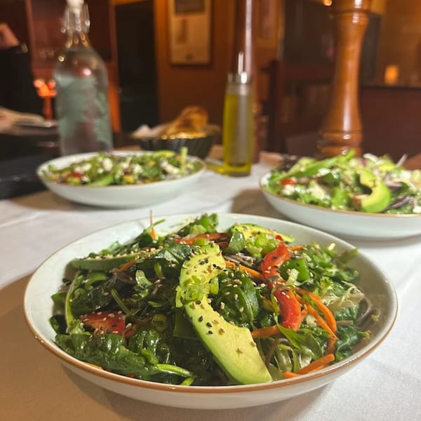Three bowls of fresh green salad with avocado slices and sesame seeds on a white tablecloth.