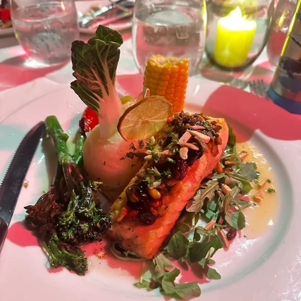 A salmon dish with bok choy, corn, broccoli, and a lime slice on a white plate.