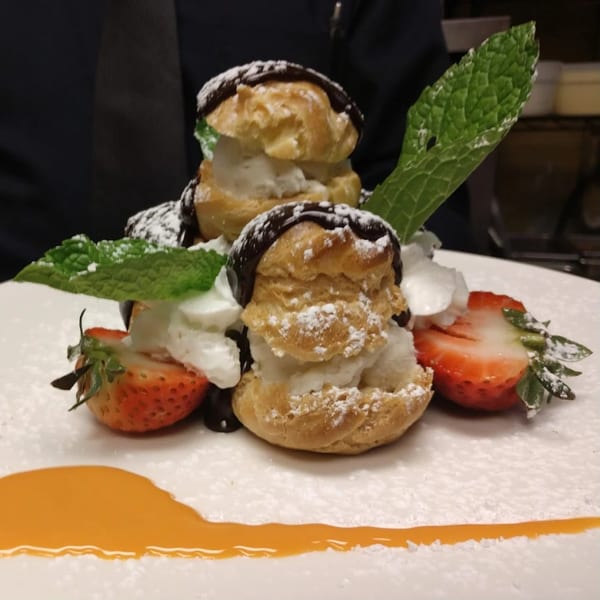 Two cream puffs with chocolate, powdered sugar, mint, and strawberries on a white plate.