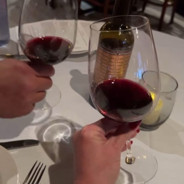 Two hands holding glasses of red wine, with a wine bottle and candle on a white table.