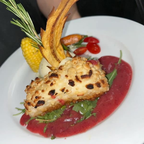 French dish with a crusted main, red sauce, corn, cherry tomatoes, and rosemary garnish.