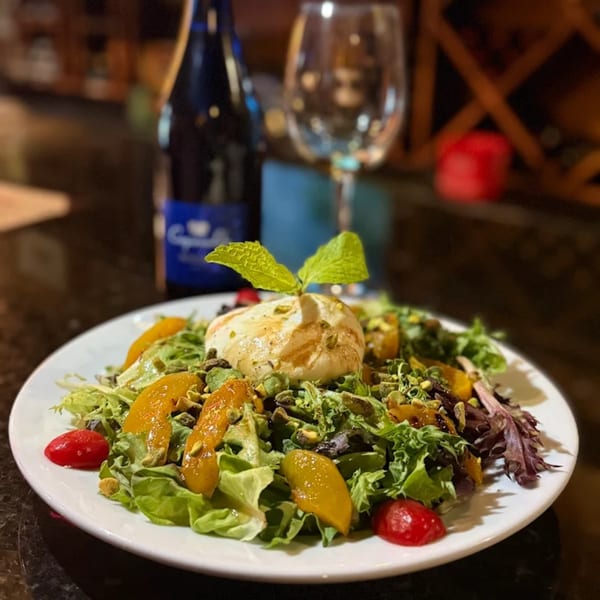 A fresh La Brochette Bistro salad with burrata, peaches, pistachios, and cherry tomatoes, with wine.